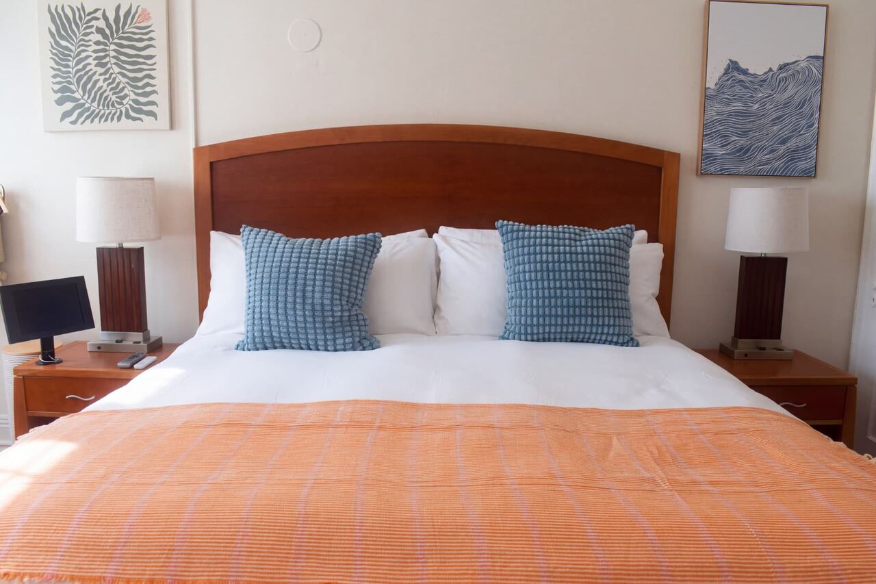 The Nook king bed with white linen, blue cushions and an orange throw between matching bedside lamps
